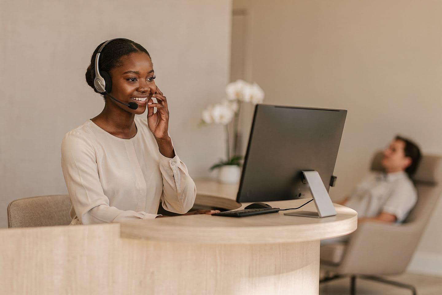 A medspa receptionist in a headset smiles on a call as a client relaxes in the lounge behind her.